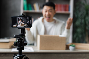 Focus on smartphone screen, happy asian male blogger unpacking gift boxes presents from companies and doing live streaming. Attractive man using smart phone camera for creating content.