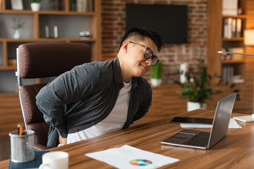 Obraz premium Unhappy middle aged asian man manager in glasses suffering from muscles back pain at table with laptop