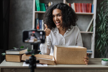 Happy female blogger unpacking gift boxes presents from companies and doing live streaming showing thumb up. Multinational woman using smart phone camera for creating content.