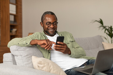 Glad middle aged african american man in glasses reads message on phone, watches video surfing in...