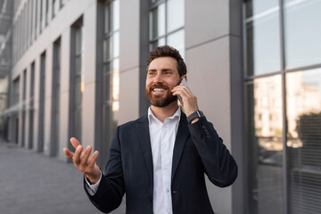 Glad young european man ceo manager with beard calls by smartphone, talks, gesticulates near office building