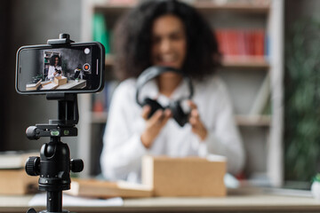 Focus on smart phone screen, woman recording video on phone camera while unpacking box with new wireless headphones. Female influencer sharing with subscribers her positive feedback about new order.