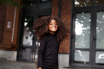 Authentic portrait African American child girl smiling and looking camera, walking city street. Beautiful fashion model kid with curly hair
