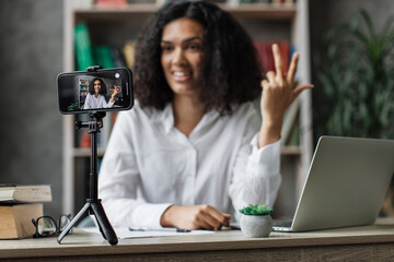 Focus on screen, charming multinational woman in white shirt talking and gesturing while recording video on modern phone on tripod. Female blogger sitting at home and doing live stream.