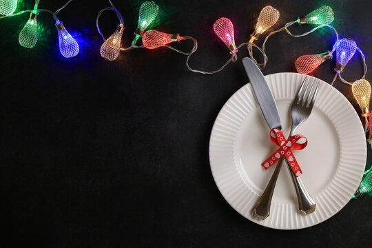 A white ribbed plate, cutlery on it, a red festive ribbon with hearts, Christmas lights, a dark background, copy space