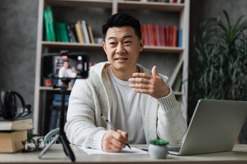 Focus on screen, smiling asian man talking and gesturing in front of modern smartphone camera on tripod, writing report while sitting on background of modern home.