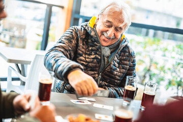 Cheerful group of seniors having fun playing card games at bar table - Old people enjoying free...
