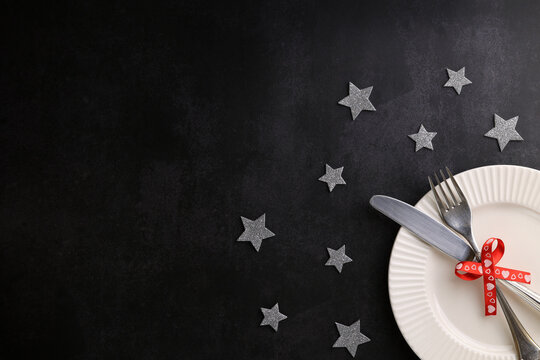A white ribbed plate, a knife and a fork on it, a red festive ribbon with hearts, gray shiny stars around, a dark background, copy space