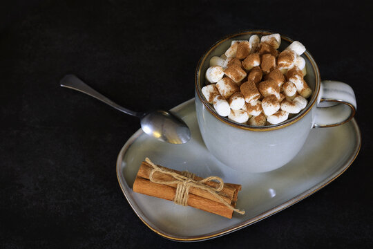 A cup of hot beverage with marshmallow and cinnamon, cinnamon sticks tied with rope, tea spoon, dark background