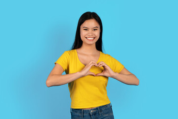 Happy pretty young korean lady in yellow t-shirt show heart sign with hands on chest, looking at camera