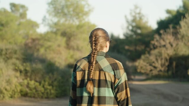 A girl with long hair and a scythe walks through the forest at sunset. She's wearing a plaid shirt, the camera follows from behind.