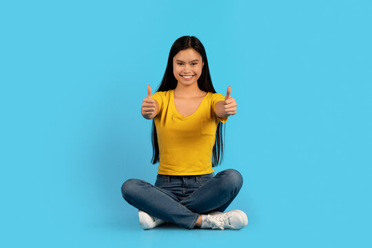 Cheerful millennial korean lady student in yellow t-shirt sitting on floor, show thumbs up, recommend order