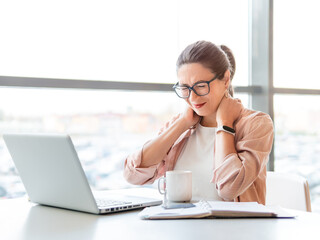 Woman winces from pain in her neck. Female office employee tries to stretch stiff neck muscles while working with laptop. Modern office at co-working center. Workplace for freelancers or students.