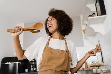 African american housewife in apron singing with spatulas for cooking microphone on the kitchen....
