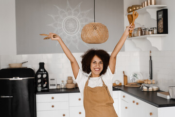 African american woman is dancing on the kitchen and raising hands up. African girl cook in apron dancing on white kitchen. Advertising for cooking courses and food workshops.