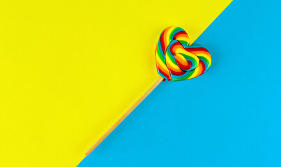 Rainbow colored heart shaped lollipop on yellow and blue background