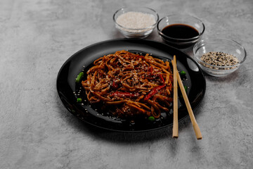 Spicy udon with chicken on a black plate with spices and soy sauce in glass bowls.