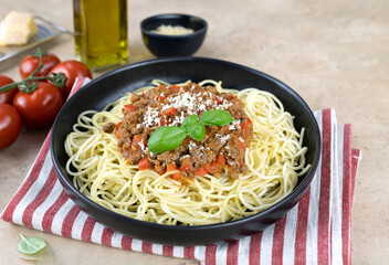 Spaghetti bolognese on a black plate with fresh basil, tomatoes, olive oil and parmesan cheese on a beige background. Italian Cuisine.