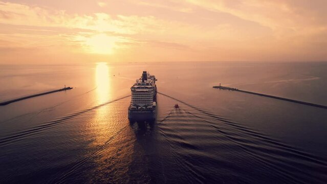 4k drone footage cruise liner at sunset in the sea.Liner slowly floats into the sea between two lighthouses,on the endless sea horizon a yellow pink sunset.Sun reflects in water.Golden hour of nature.