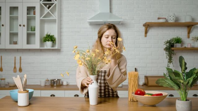A housewife creates coziness at home using flowers. A woman sniffs flowers in a vase and enjoys the smell and thinks about something good. Modern kitchen interior.