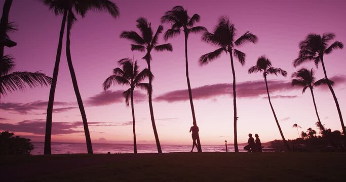 Beach vacation travel sunset background with woman running and people enjoying view of perfect pink colorful sunset with palm trees. Running woman in silhouette jogging at beach sunrise in SLOW MOTION