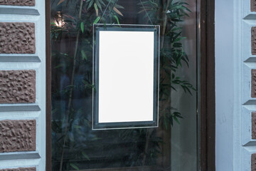 Empty white poster in glass frame fixed on window of street cafe or shop. Blank signboard for...