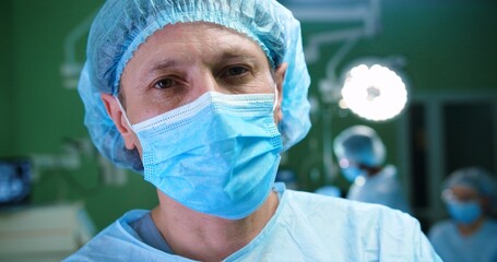 Close up portrait of Caucasian joyful middle-aged male doctor surgeon in medical uniform putting off mask and smiling to camera after surgery. Team of surgeons in operating room on background