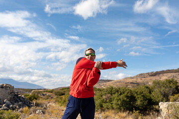 Fototapeta na wymiar A man, dressed in a tracksuit and sunglasses, stretches his back, arms and muscles during a walk in the countryside on a sunny day