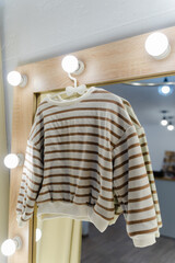 Jumper hangs on a hanger on a mirror with light bulbs. Fitting room with a large beautiful mirror with light bulbs. Children's sweater with stripes. Beige sweater with brown stripes. Vertical.