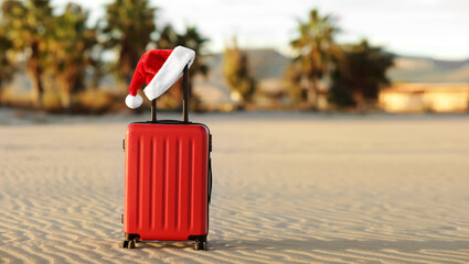 Red suitcase with Santa Claus red hat on handle with tropical sand beach, palms, mountains and sea background. Travel concept for the Christmas holidays and New Year. Place for text. Discounted trips