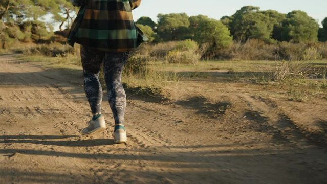 A girl in leggings and a plaid shirt runs along a sandy road in the forest at sunset. the rear view is slow-motion