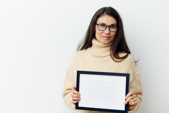  happy, neat woman in a beige turtleneck sweater and black-rimmed glasses, standing on a light background holding a white frame with a black frame in her hands. Horizontal photo for advertising text
