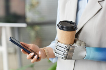 Closeup image of woman with prosthetic arm holding cup of take out coffee and checking notifications on smartphone
