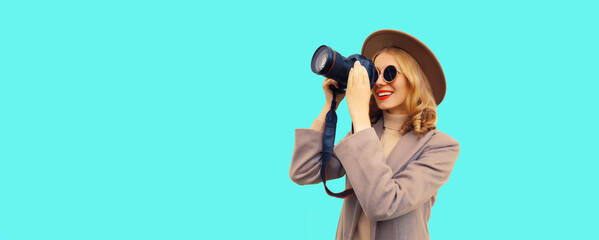 Portrait of stylish happy smiling woman photographer with digital camera taking picture wearing round hat, brown coat on blue background, blank copy space for advertising text