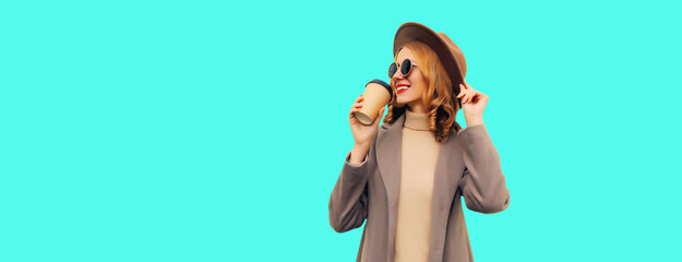 Portrait of stylish beautiful young woman model with cup of coffee wearing brown round hat and coat on blue background