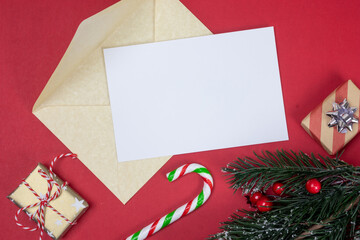 Christmas letter mock up with golden envelope and blank empty white paper page with copy space top view. Fir tree branch, holly, gold colour presents and red and green candy cane.
