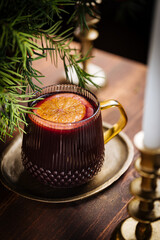 Mulled wine with cinnamon and orange served in festive Christmas decoration on wooden board. Party and New Year atmosphere. Christmas tree in background, a mug with Christmas motive. 