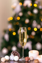 New year glass of sparkling wine, white christmas decorations and candle on a table in interior in front of green christmas tree decorated with yellow bokeh lights. Merry Christmas and happy New year.