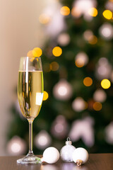 New year champagne in a glass and white christmas toys on a wooden desk in front of christmas tree decorated with yellow garland lights. Waiting for winter holidays. New Year atmosphere.