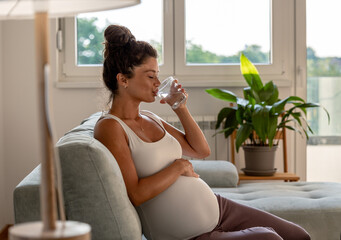Pregnant woman drinking water on sofa at home - 549840954