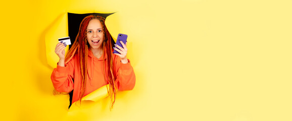 Young woman with dreadlocks, mobile phone, credit card, sticking out of hole of on yellow background with space for text. Female browsing smartphone, entering card details, paying internet purchase.