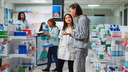 Asian worker explaining pharmacy leaflet in medical retail store, giving advice to find prescription treatment and pharmaceutical products. Young man looking at vitamins and medicaments on shelves.