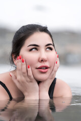 Portrait of woman embracing face with her palms, playfully looking up. Plus size brunette model in black swimming costume lying down in water of outdoor pool at balneotherapy spa, hot springs resort - obrazy, fototapety, plakaty