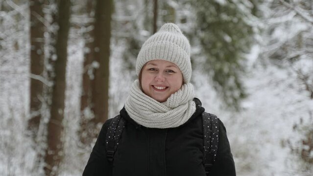 Portrait of young overweight woman in warm clothes in winter snowy forest. Lady in beige hat, snood with walking backpack on shoulders smiles and looks at camera. Woman rejoices in the winter season.