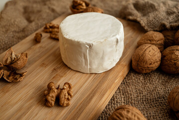 Composition of a round head of cheese with white mold and walnuts on a bamboo board on a background of burlap