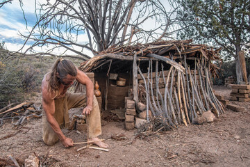 Man making friction fire in front of shelter - obrazy, fototapety, plakaty