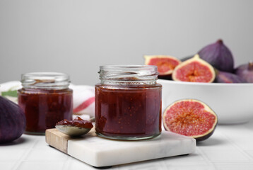Glass jars of tasty sweet fig jam and fruits on white tiled table