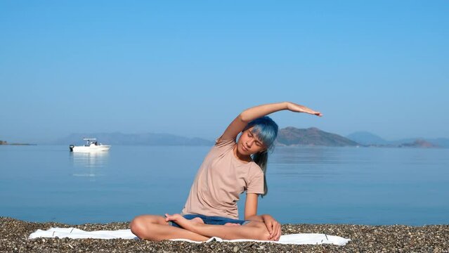 Teen stretching exercises on shore. A view of active cute girl stretching her body in lotus pose on the empty shore in the morning.