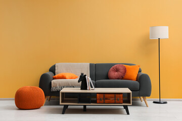 Stylish grey sofa with colorful pillows, wooden table and lamp near pale orange wall indoors....