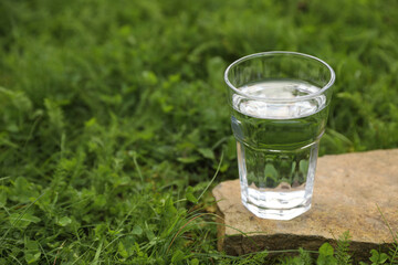 Glass of fresh water on stone in green grass outdoors. Space for text